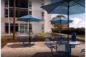 Outdoor patio area with round tables and blue chairs under large blue umbrellas, situated in front of a multi-story building with balconies and windows. There are some small trees and landscaped areas around the patio.