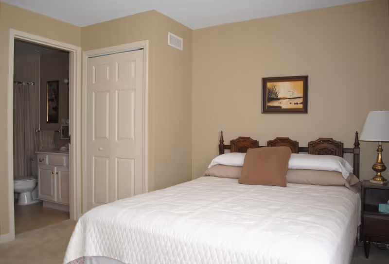 A bedroom with a neatly made bed featuring a white quilt and brown pillows. The headboard is wooden with intricate designs. To the right of the bed is a dark wooden nightstand with a brass lamp and a box of tissues. On the wall above the bed hangs a framed painting of a serene lakeside scene with trees and birds. To the left, an open door reveals a bathroom with a toilet, sink, and shower curtain. The walls are painted beige.