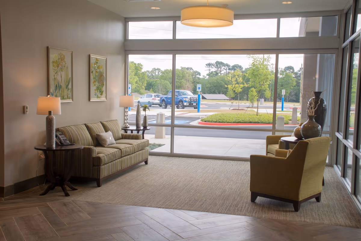 A bright and cozy seating area inside a facility with a large window showing a parking lot and greenery outside. The room features a striped sofa with a pillow, two side tables with lamps and decorative vases, two armchairs, and framed floral artwork on the wall.