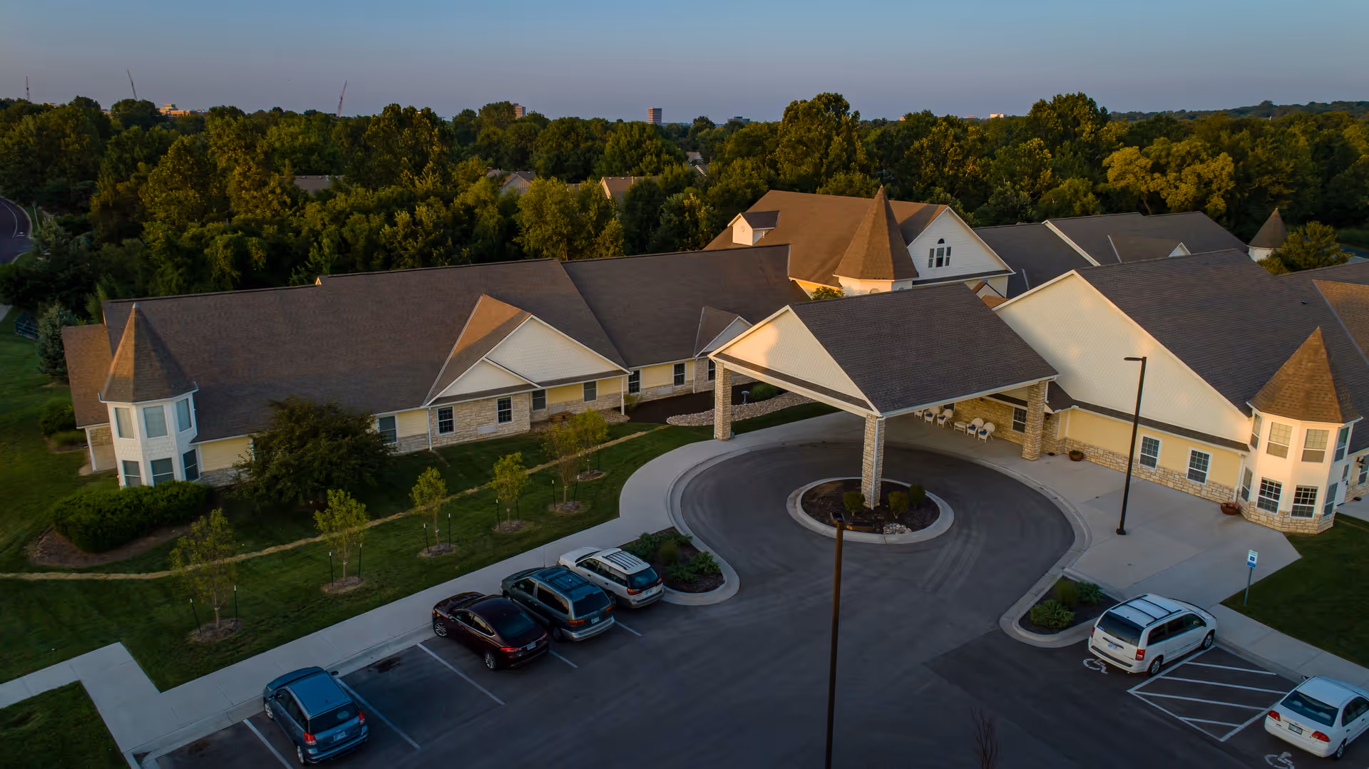 Aerial view of Homestead Assisted Living & Memory Care of Overland Park, showing a large building with multiple peaked roofs and turrets, surrounded by trees and greenery. The facility has a circular driveway with a covered entrance and several parked cars in the parking lot.