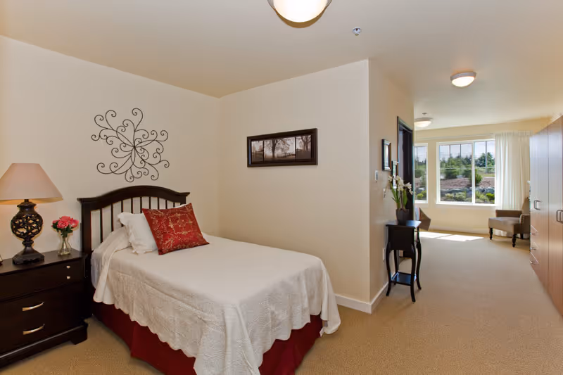 A well-lit senior living bedroom with a single bed covered in a white quilt and a red decorative pillow. There is a dark wooden nightstand with a lamp and a small vase of flowers next to the bed. The room has beige walls with a decorative metal wall art above the bed and a framed picture on the adjacent wall. A small black table with a plant is placed near a hallway leading to a bright sitting area with a large window and two chairs. A wooden wardrobe is visible on the right side of the room.