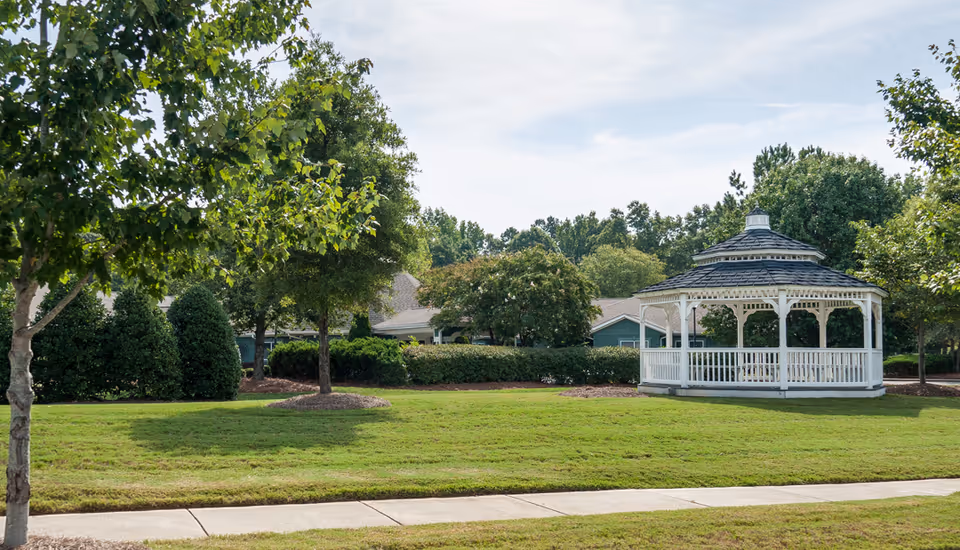 A well-maintained outdoor area at Mint Hill Senior Living featuring a white gazebo surrounded by green grass, trees, and shrubs under a partly cloudy sky.