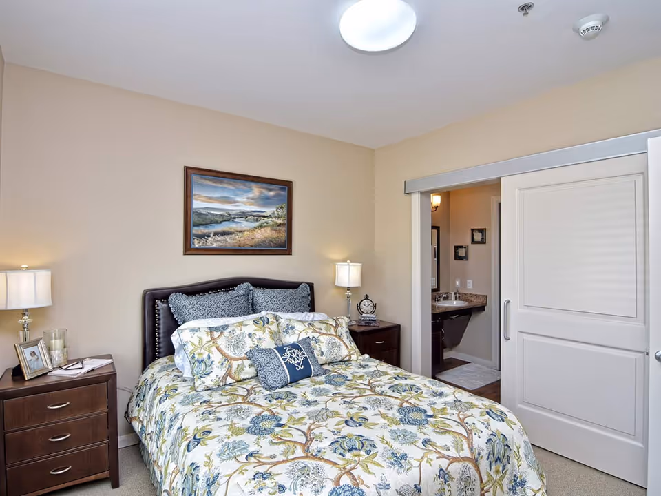 A cozy bedroom with a floral patterned bedspread on a double bed, two bedside tables each with a lamp, and a framed landscape painting on the beige wall. A sliding door is partially open, revealing a bathroom with a sink and mirror inside.