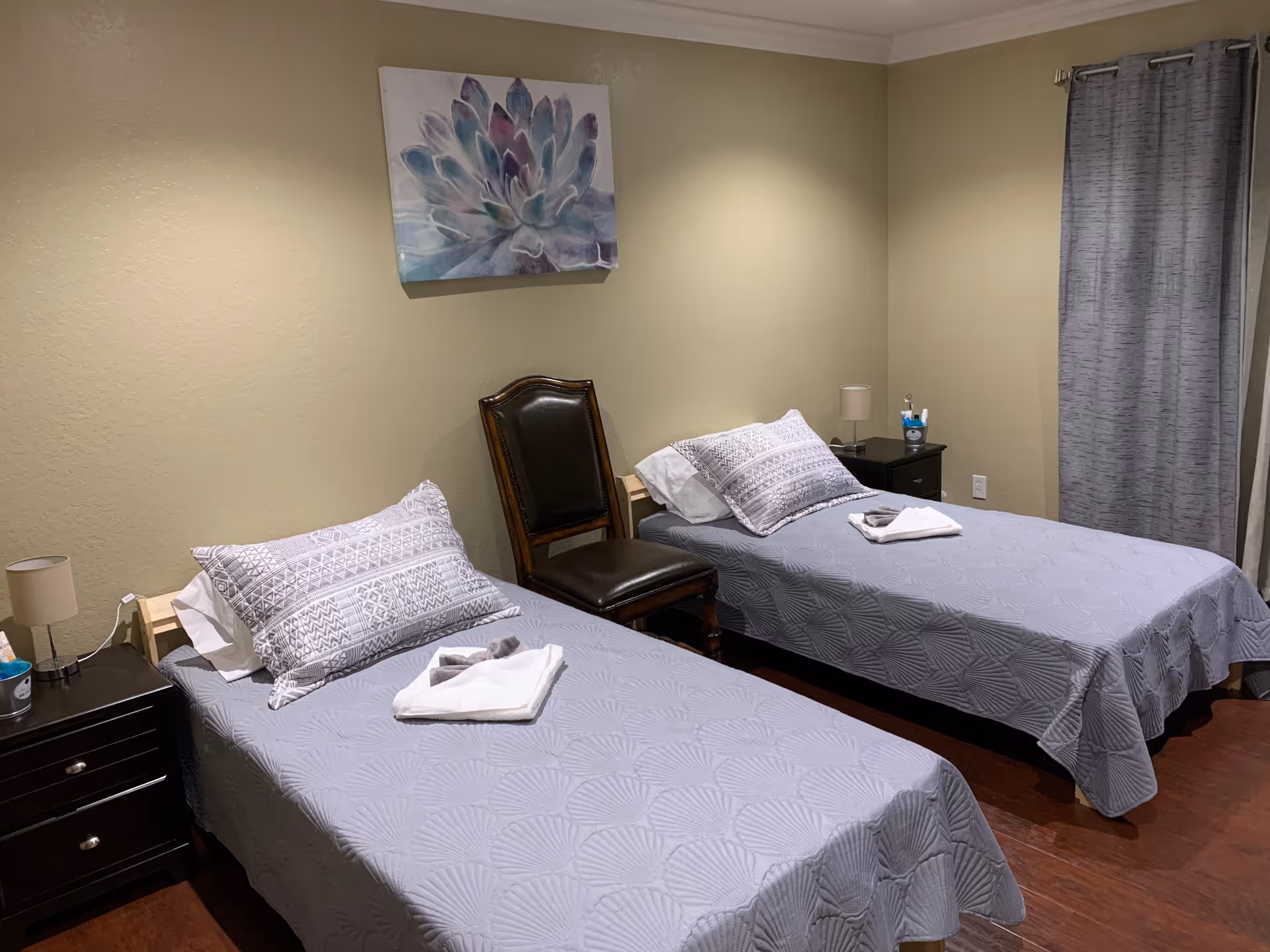 A bedroom with two single beds covered in gray patterned bedspreads, each with a pillow and a folded towel on top. There is a wooden chair with a dark cushion placed between the beds. On either side of the beds are black nightstands with small lamps and some items on top. A large painting of a succulent plant hangs on the beige wall above the beds, and gray curtains cover a window on the right side of the room.