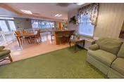 A spacious common area in a senior living facility featuring a green carpeted seating area with a cushioned armchair and a small side table. Beyond the seating area, there is a dining space with wooden tables and chairs on a light wood floor, large windows with floral curtains allowing natural light to fill the room.
