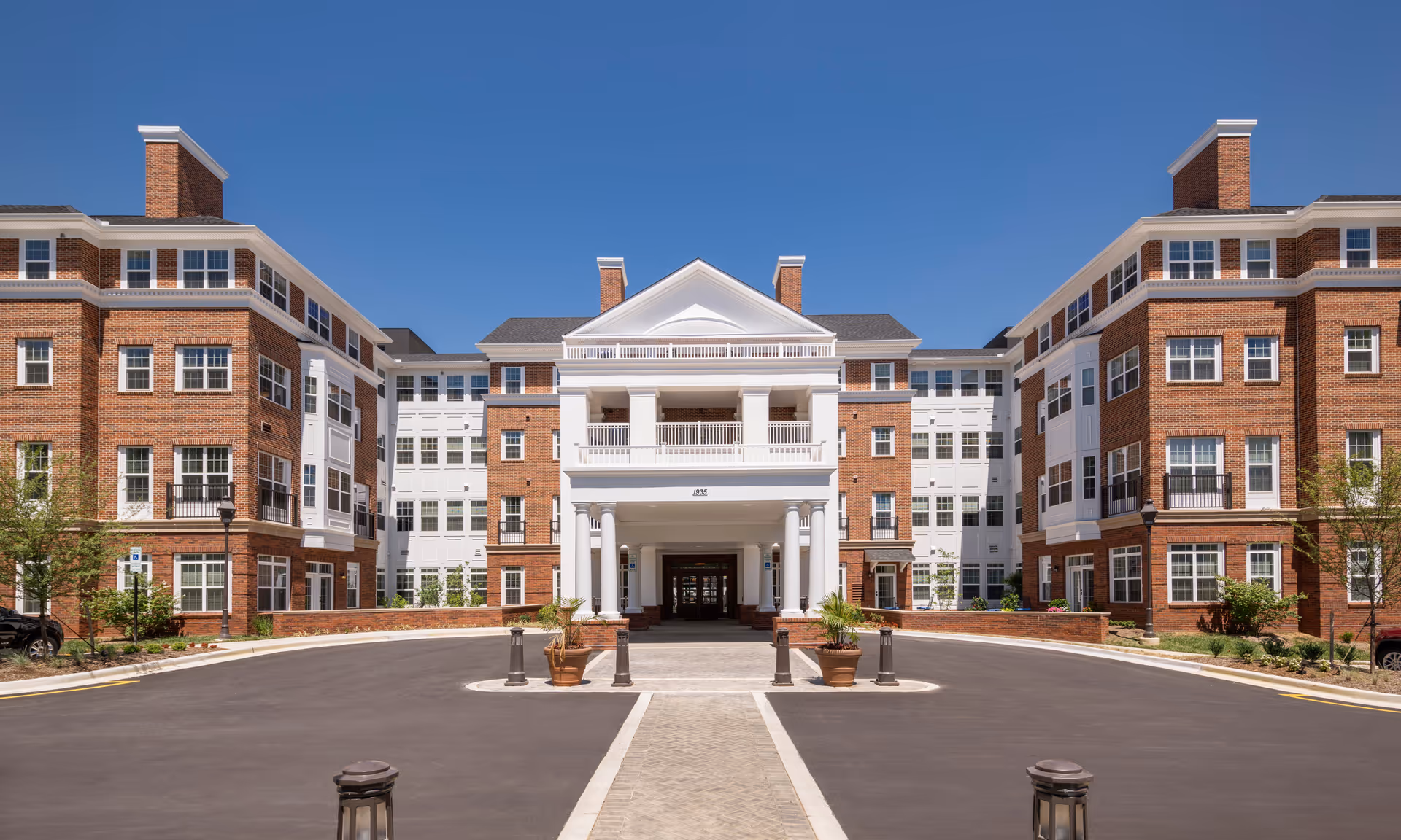 Front exterior view of a large senior living facility building with red brick and white accents, featuring multiple windows and a central entrance with white columns under a clear blue sky.