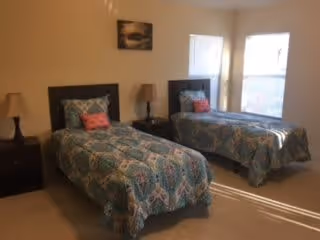 A bedroom with two twin beds, each with patterned blue and white bedding and a pink decorative pillow. There are two bedside tables, each with a lamp. A framed picture hangs on the wall above one bed, and two windows allow natural light into the room.