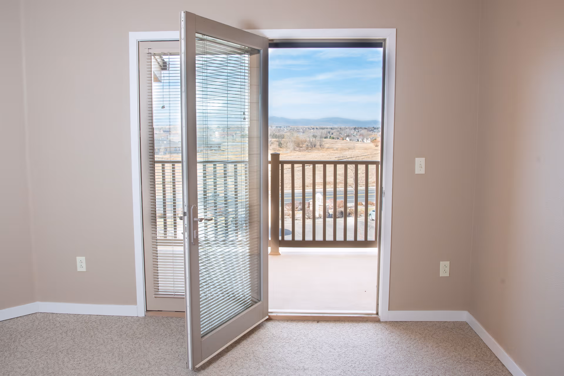 An empty room with beige walls and carpeted floor featuring a glass door with blinds that opens to a balcony overlooking a scenic outdoor view with clear skies and distant mountains.
