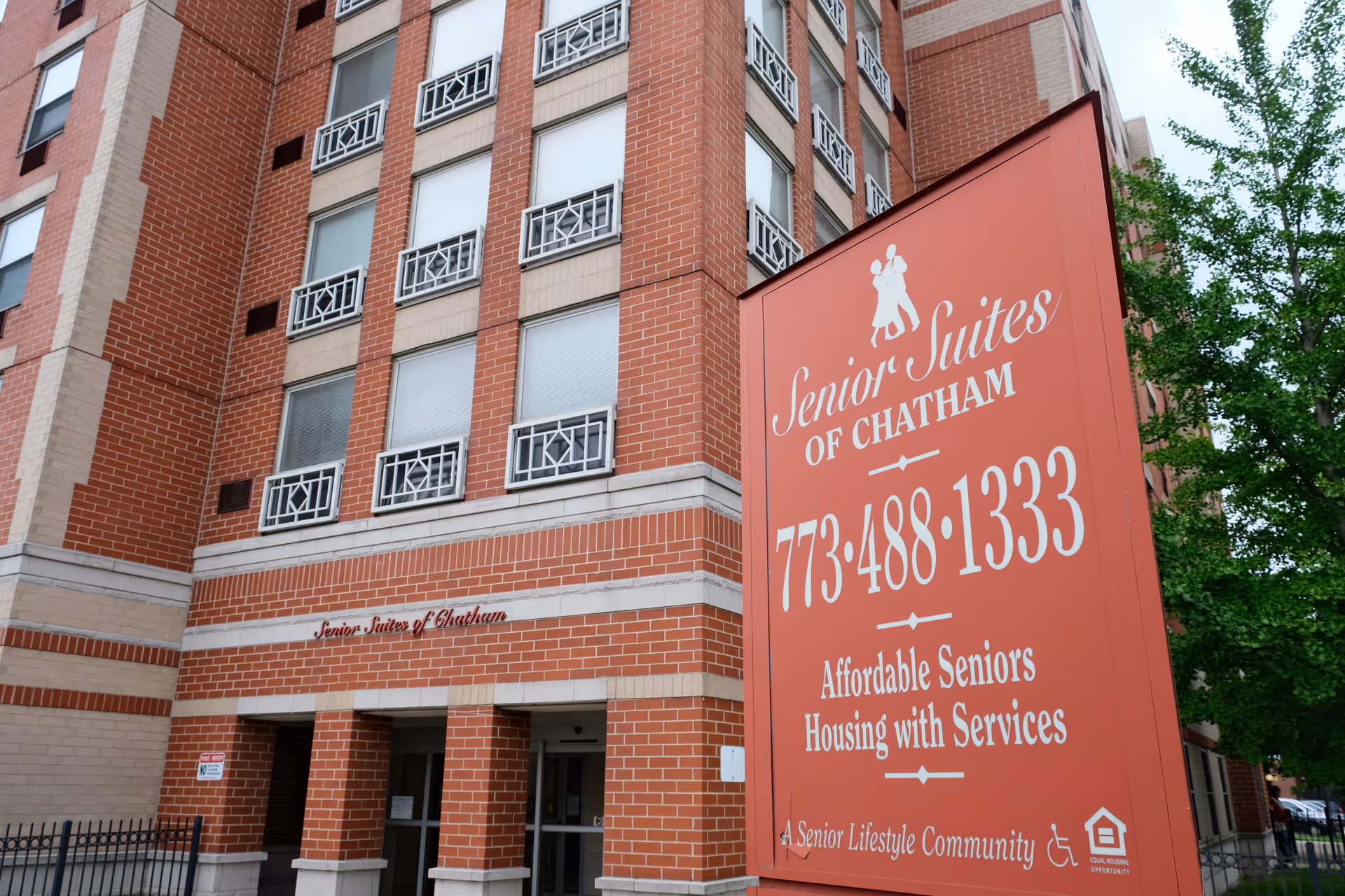 Exterior view of a multi-story brick building with windows and decorative metal railings. In front of the building is a large red sign that reads 'Senior Suites of Chatham' with a phone number, and information about affordable seniors housing with services. There are trees visible to the right side of the image.