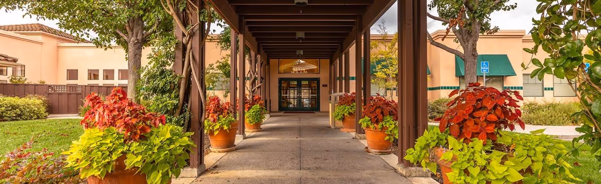 Covered walkway with wooden beams and potted plants on both sides leading to the entrance of a building with glass doors. The building has beige walls, green awnings, and trees around the pathway.