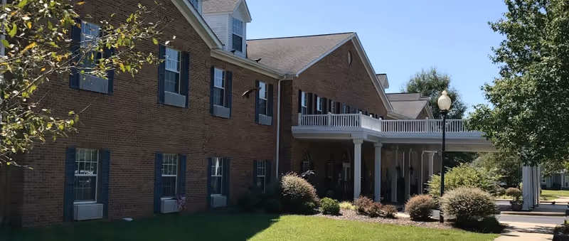 Brick two-story retirement community building with a covered columned entrance, landscaped lawn, and a clear blue sky.