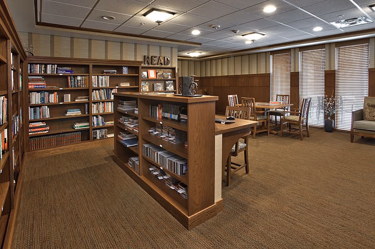 A cozy library room with wooden bookshelves filled with books and CDs, a wooden table with chairs, and large windows with blinds letting in natural light. The word 'READ' is displayed on top of one bookshelf.