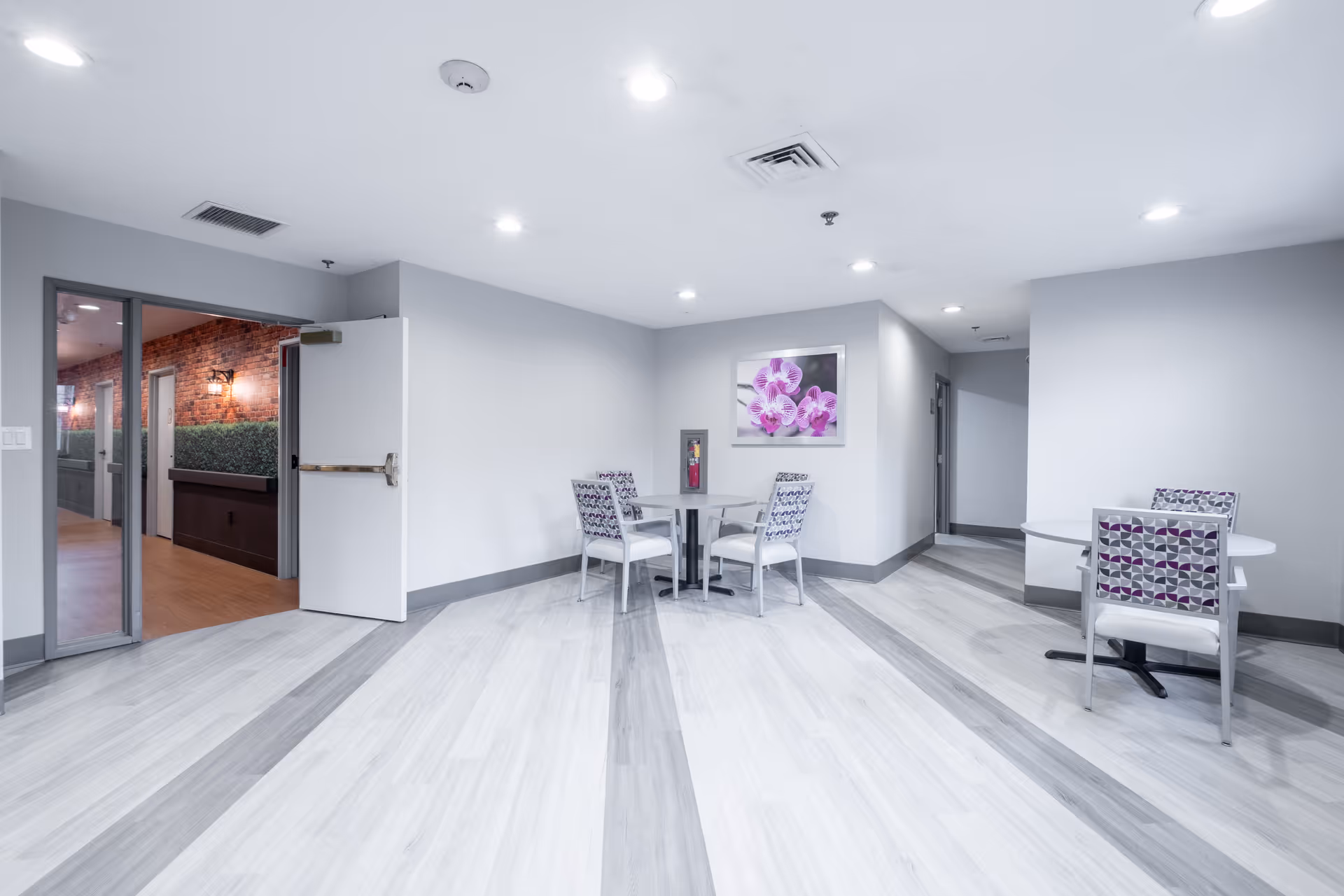A bright interior room with light gray walls and flooring featuring darker gray stripes. There are two small round tables, each with two patterned chairs. A framed picture of purple orchids hangs on the wall. An open door leads to a hallway with brick walls and greenery. The ceiling has multiple recessed lights and ventilation vents.