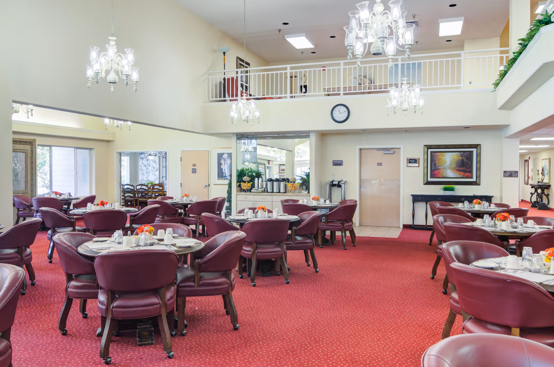 Spacious assisted-living dining room with round tables set for meals, burgundy chairs, red carpet and chandeliers.