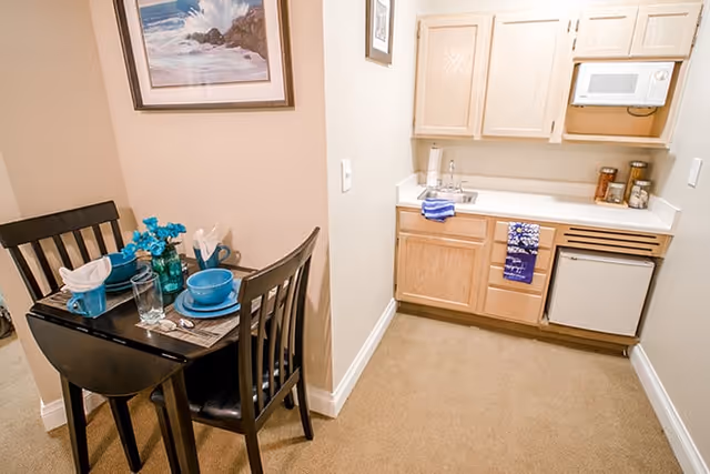 Small kitchenette with light wood cabinets, microwave, mini-fridge and a small two-person dining table set with teal dishware.