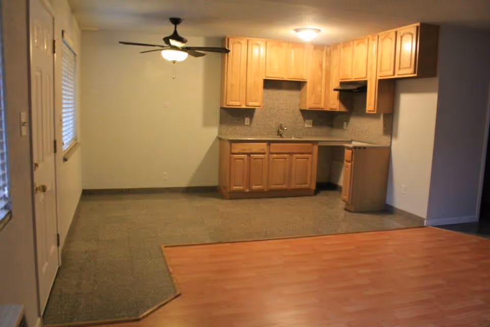 Open-plan interior showing a small kitchenette with wooden cabinets and sink, a ceiling fan, and flooring that transitions from tile to laminate.
