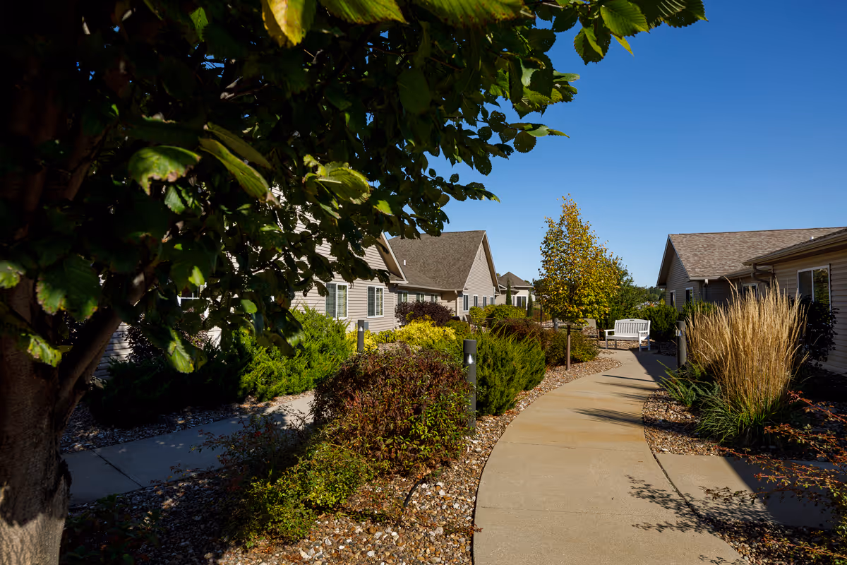A sunny outdoor pathway winding through landscaped gardens with bushes, trees, and ornamental grasses, flanked by single-story buildings with beige siding and brown roofs. A white bench is visible along the path under a clear blue sky.