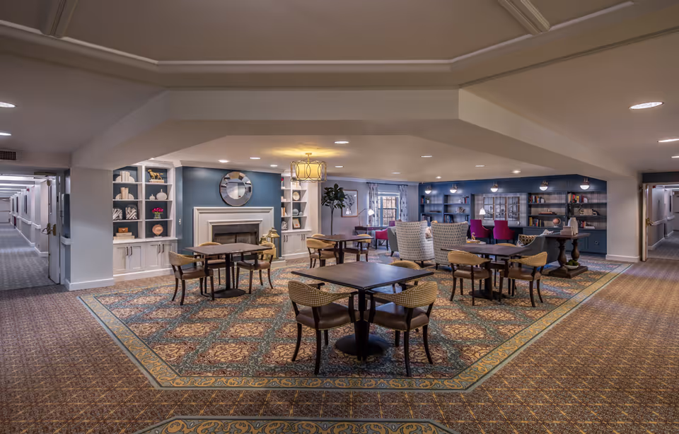 Spacious senior living common lounge with tables and chairs, a fireplace, bookshelves and a seating area.