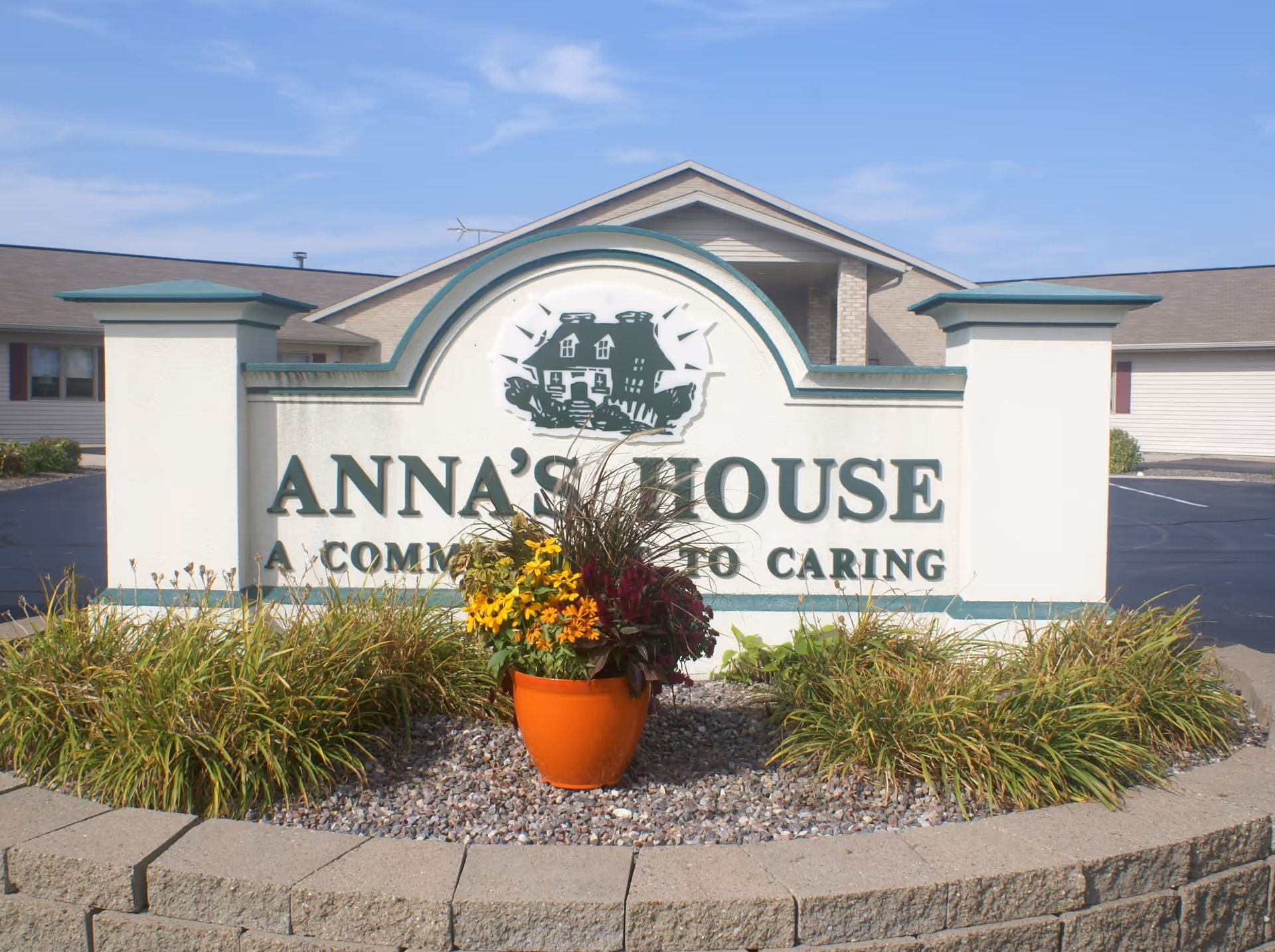 Outdoor view of a sign for Anna's House, a community dedicated to caring, with a flower pot containing yellow and dark red flowers in front of the sign. The building and parking lot are visible in the background under a partly cloudy sky.