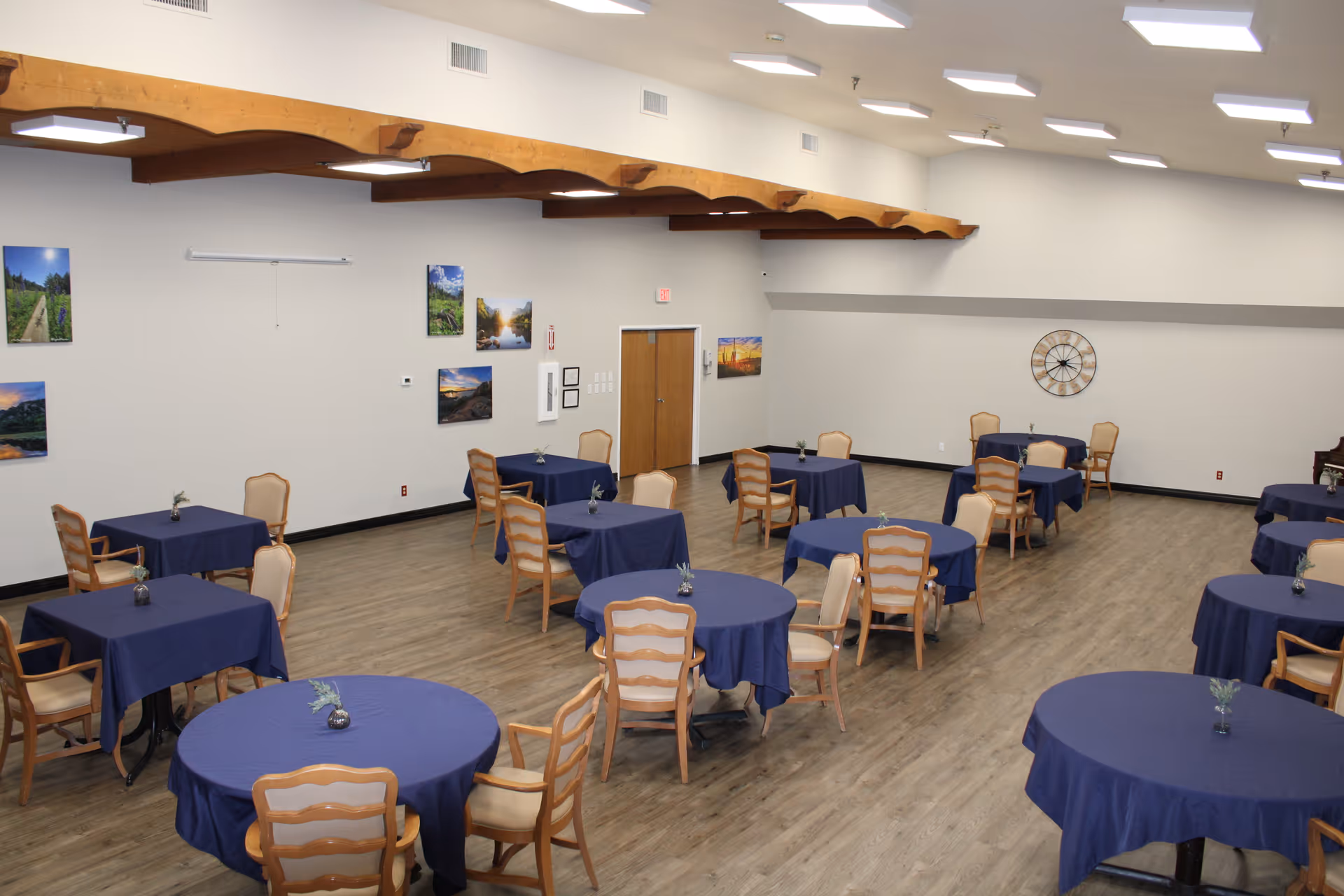 A spacious dining room with multiple round and square tables covered with blue tablecloths. Each table is surrounded by wooden chairs with light-colored cushions. The room has wooden flooring, white walls adorned with landscape pictures, and a large decorative clock on one wall. The ceiling features wooden beams and recessed lighting.