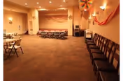 A large room with beige walls and carpeted floor, decorated with orange streamers and pinwheels. There are rows of chairs lined up along the right wall and folding tables with chairs on the left side. The room appears to be set up for a gathering or event.
