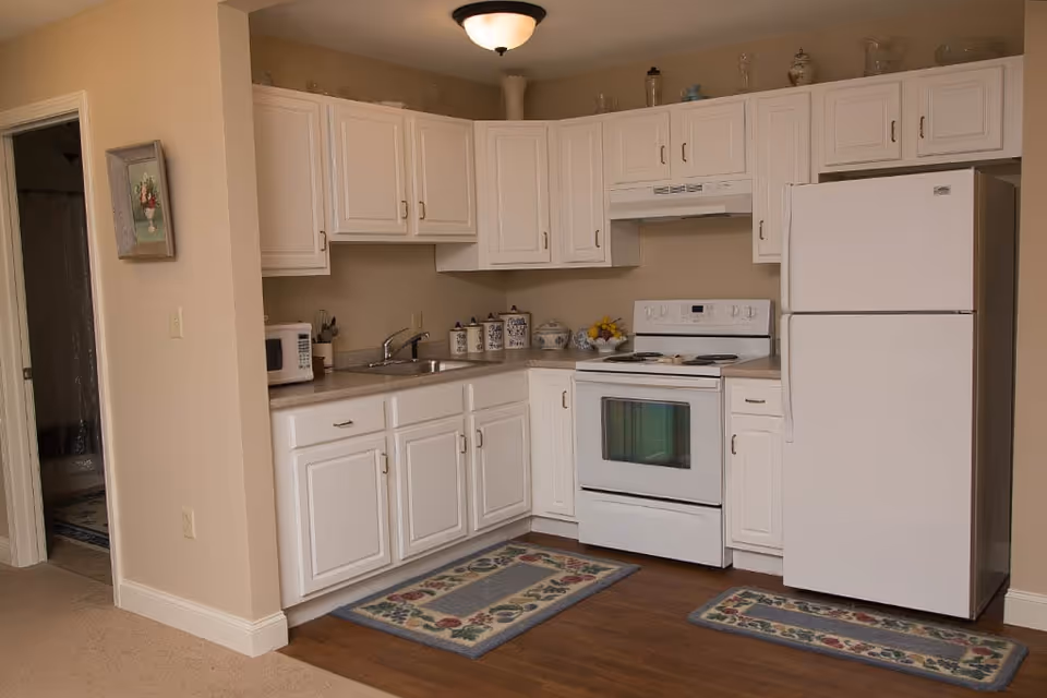 A compact kitchen with white cabinets, a white refrigerator, a white stove with an oven, a microwave on the countertop, a sink, and decorative canisters. The kitchen has beige walls and two floral-patterned rugs on the wooden floor. A doorway to a bathroom is visible on the left side.