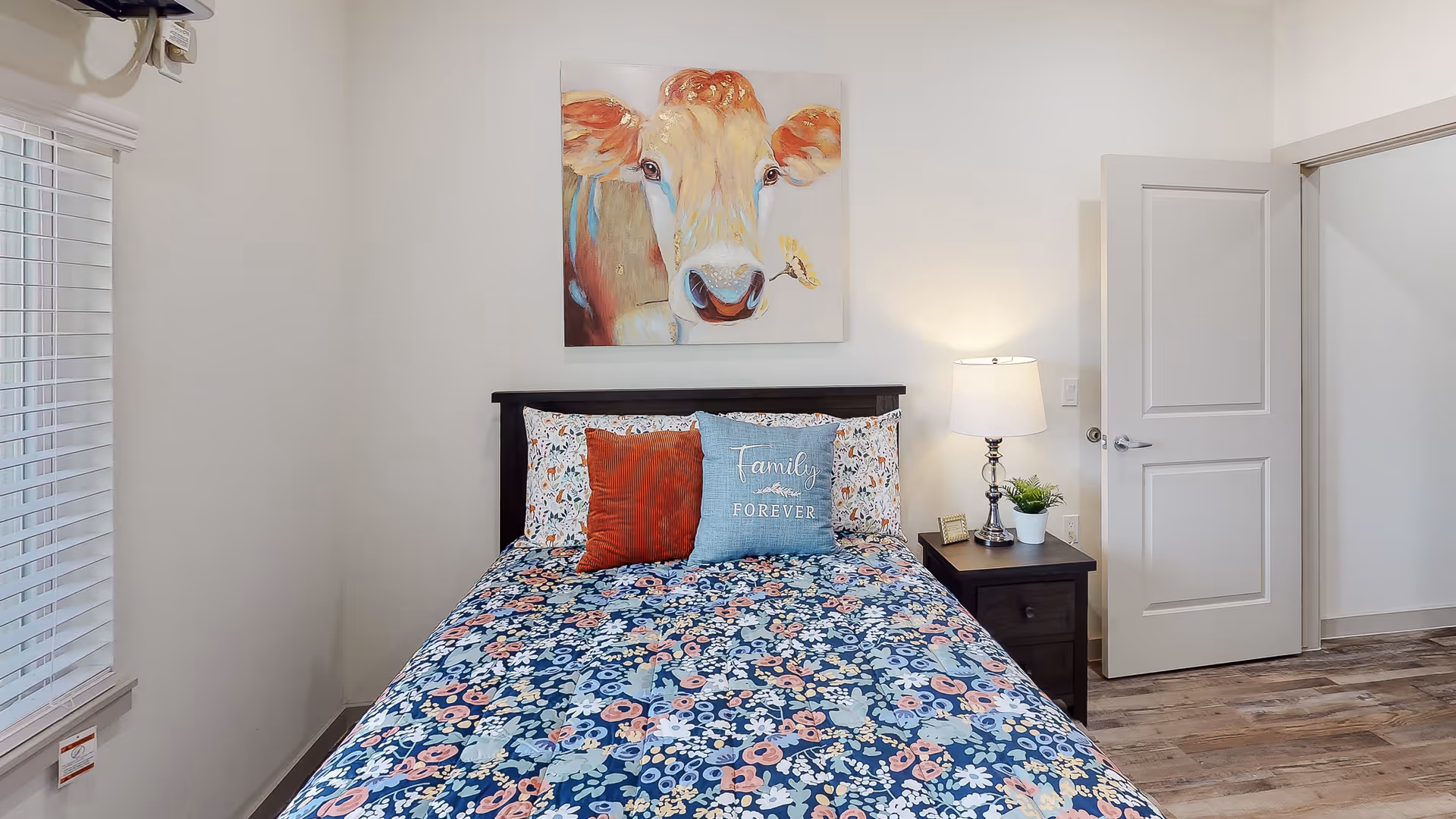 A bedroom with a bed covered in a floral patterned bedspread and two decorative pillows, one orange and one blue with the words 'Family Forever'. Above the bed hangs a colorful painting of a cow with a butterfly on its nose. To the right of the bed is a dark wooden nightstand with a lamp, a small plant, and a picture frame. The room has light-colored walls, a window with white blinds on the left, and an open door on the right showing a glimpse of another room with similar flooring.