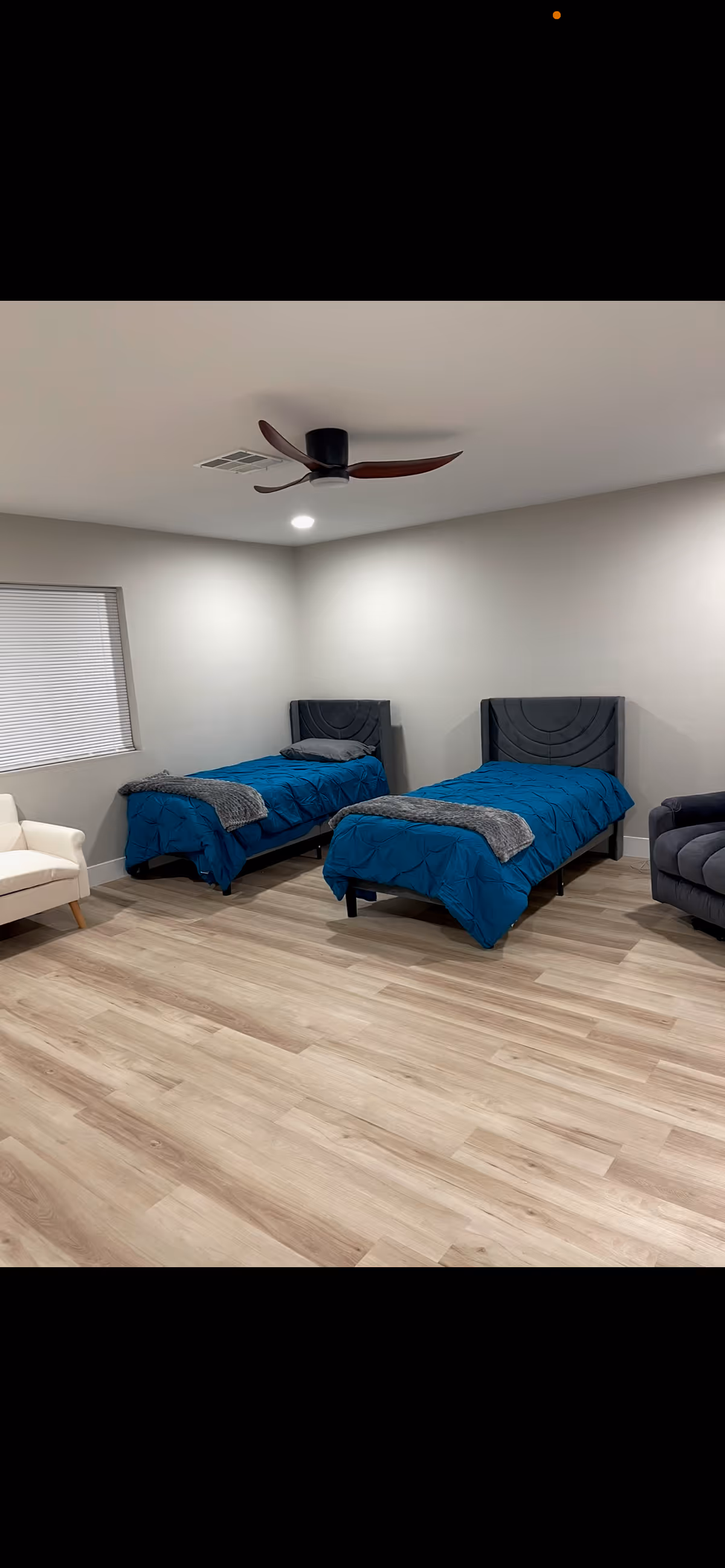 A minimal bedroom with two twin beds made with blue bedding, a ceiling fan, and seating along the walls.