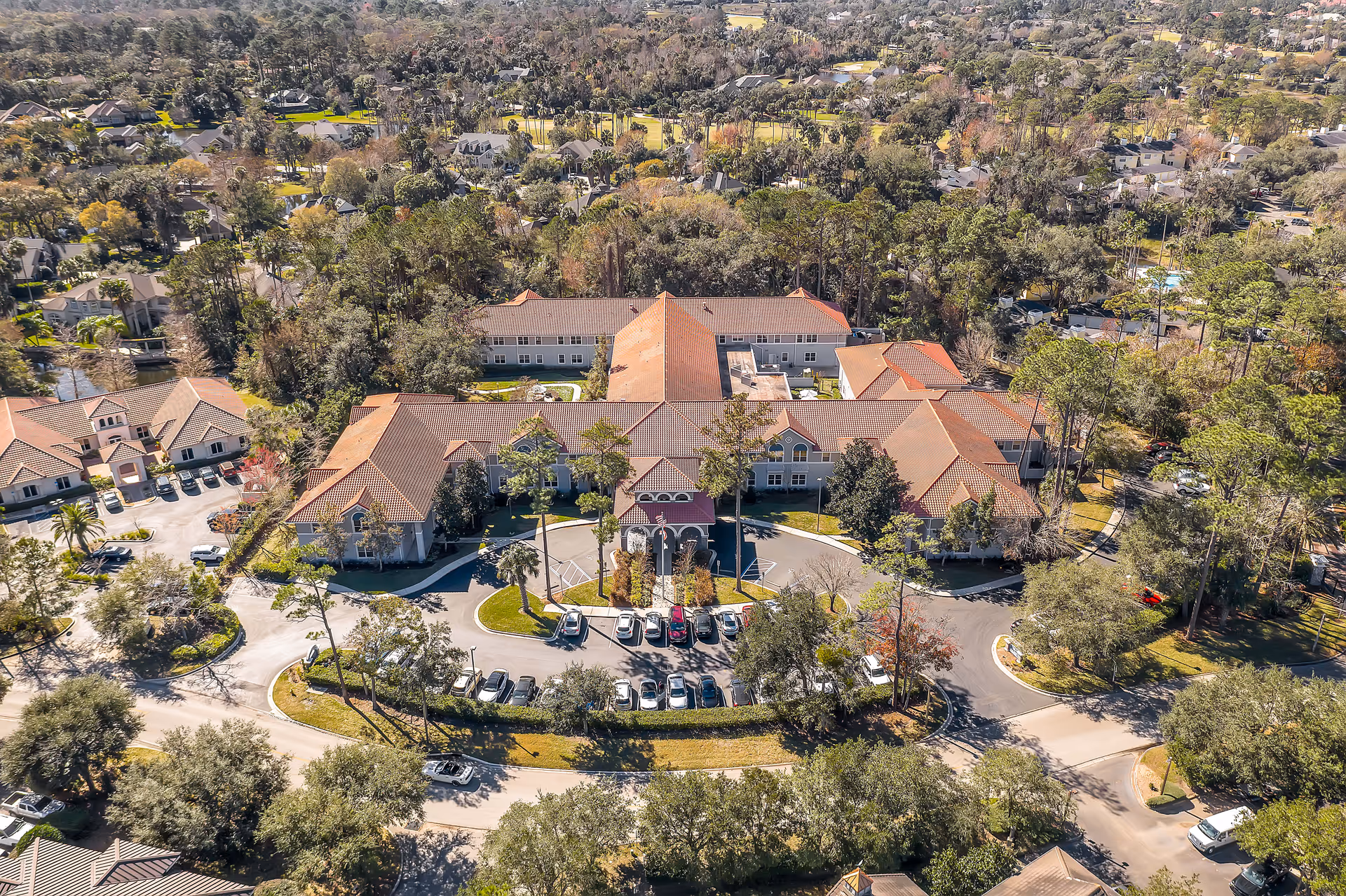 Aerial view of The Cove at Marsh Landing senior living facility surrounded by trees and residential neighborhoods, showing a large building with a red-tiled roof and a circular driveway with parked cars.