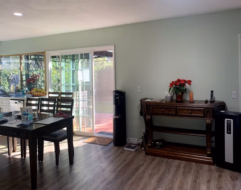 Sunlit open dining area with a wooden table and chairs by sliding glass doors and a wooden console table against a pale green wall.