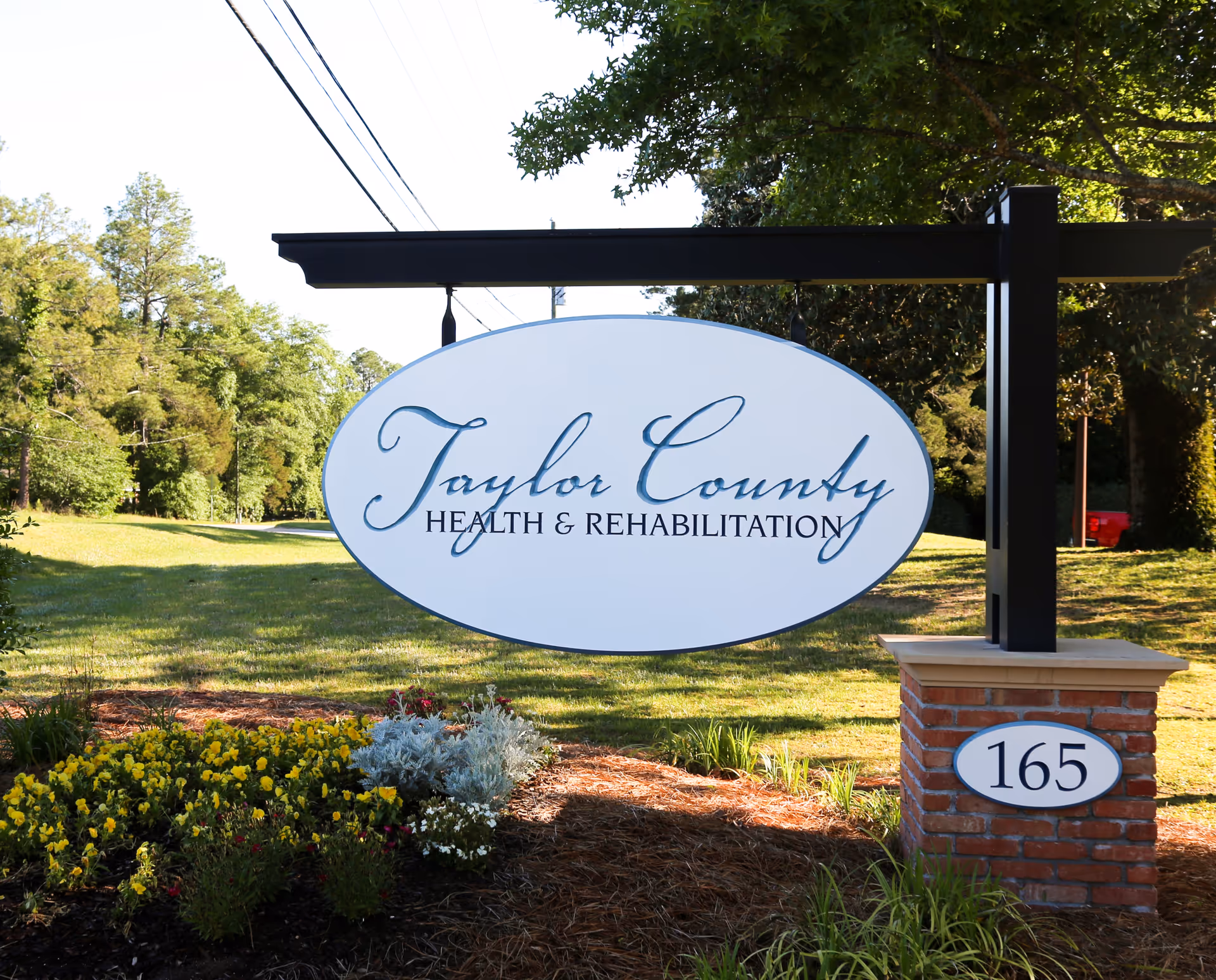 Outdoor sign for Taylor County Health & Rehabilitation mounted on a black metal frame with a brick base, surrounded by grass, flowers, and trees in the background.