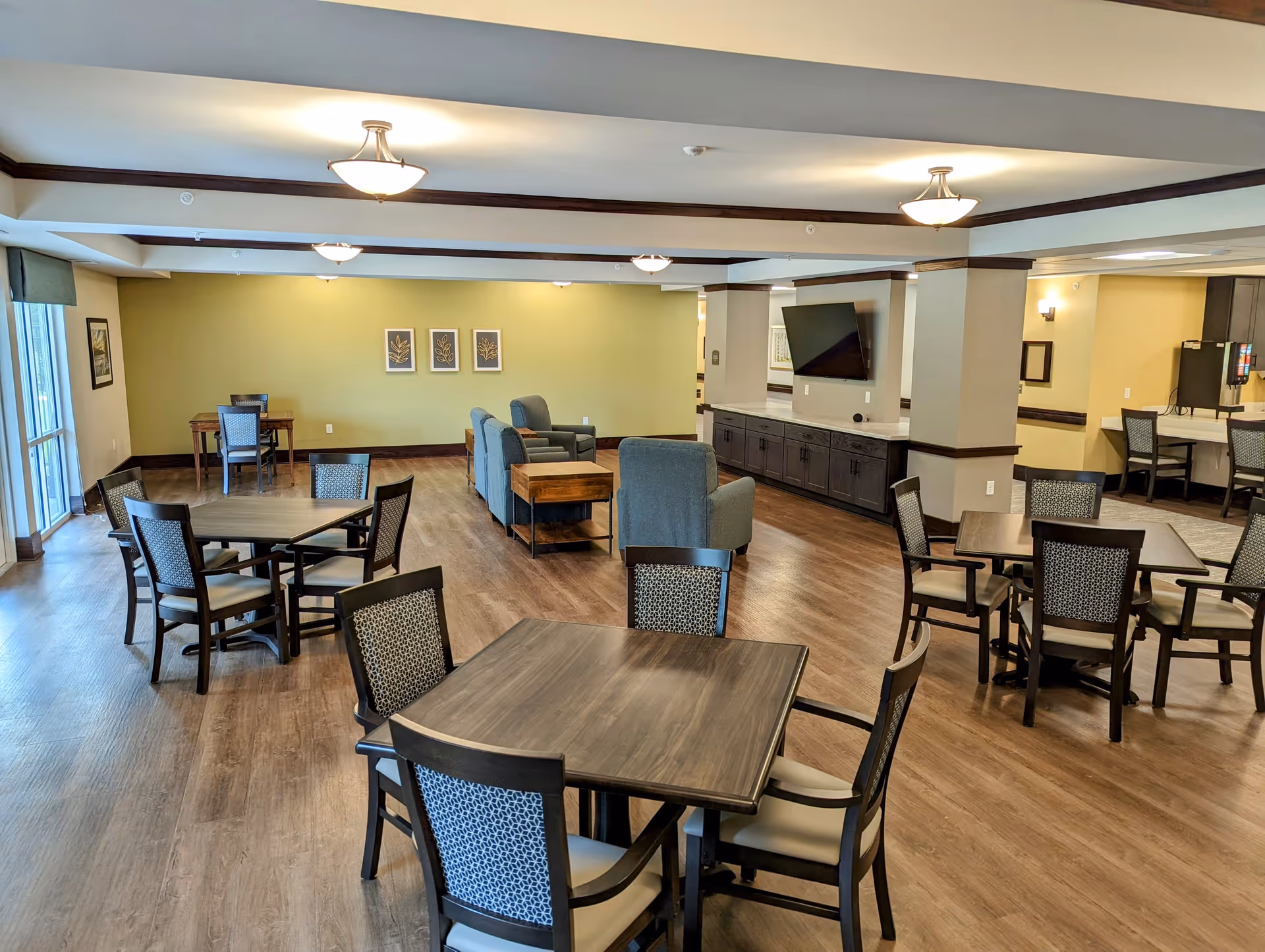A spacious senior living common area with multiple square wooden tables surrounded by chairs, a seating area with blue upholstered armchairs and wooden side tables, a wall-mounted flat screen TV, and a coffee station with chairs along a counter. The room has wood flooring, soft yellow and beige walls, and ceiling lights.
