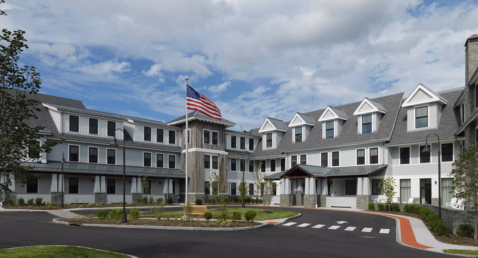 Exterior view of a large, modern senior living facility with multiple windows, a covered entrance, a circular driveway, landscaped greenery, and an American flag flying on a flagpole in front of the building under a partly cloudy sky.