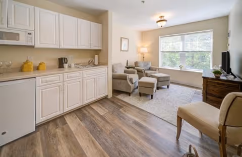 A bright and cozy living area in an assisted living facility featuring a kitchenette with white cabinets, a small refrigerator, and a microwave. The room has wood-style flooring and a seating area with two armchairs, a matching ottoman, a side table with a lamp, a large window with blinds, and a TV on a wooden stand. There is also a beige chair partially visible in the foreground.