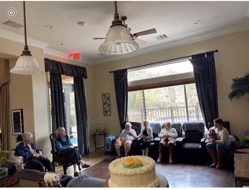 A group of elderly women sitting in a living room area with large windows and dark curtains. One woman is playing a guitar while the others listen. There is a cake with floral decoration in the foreground and pendant lights hanging from the ceiling.