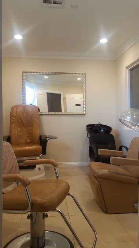 Interior view of a salon area with three tan and black salon chairs, a large mirror on the wall, and a hair washing station with a black basin. The room has beige walls and tiled floor with ceiling lights.