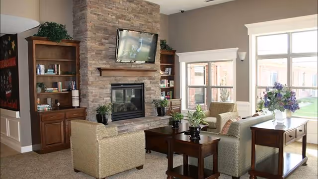 A cozy living room area with two armchairs and a sofa arranged around a wooden coffee table and side tables. The room features a stone fireplace with a mounted flat-screen TV above it, flanked by two wooden bookshelves filled with books and decorative items. Large windows allow natural light to fill the space, and there are plants and flowers adding greenery to the room.
