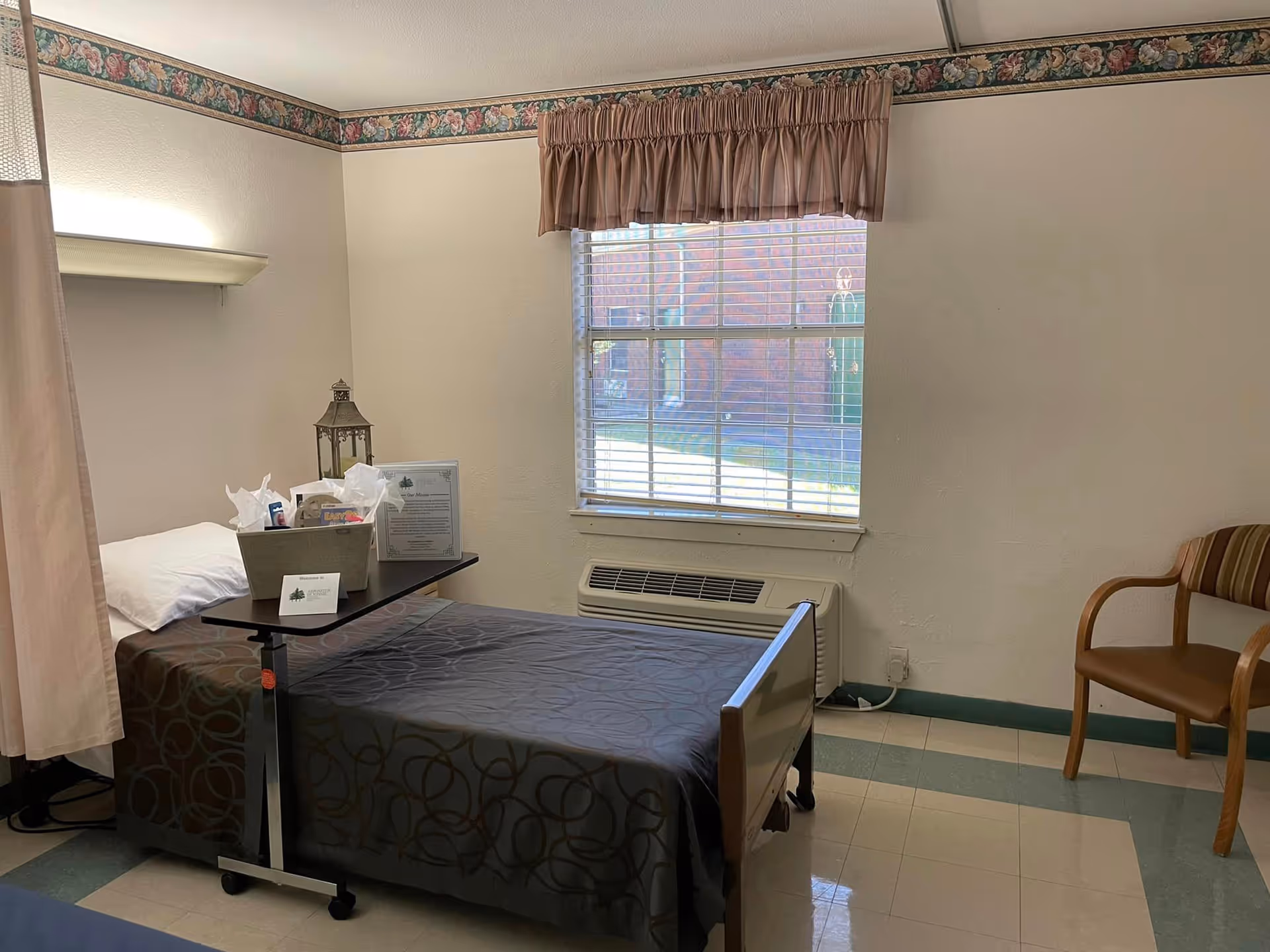 A simple nursing facility bedroom with a single bed covered in a dark patterned blanket, a pillow, and an overbed table holding a basket with tissues and other items. There is a window with blinds and a brown valance, a wooden chair with striped upholstery, and a wall-mounted air conditioning unit below the window. The walls are light-colored with a floral border near the ceiling.
