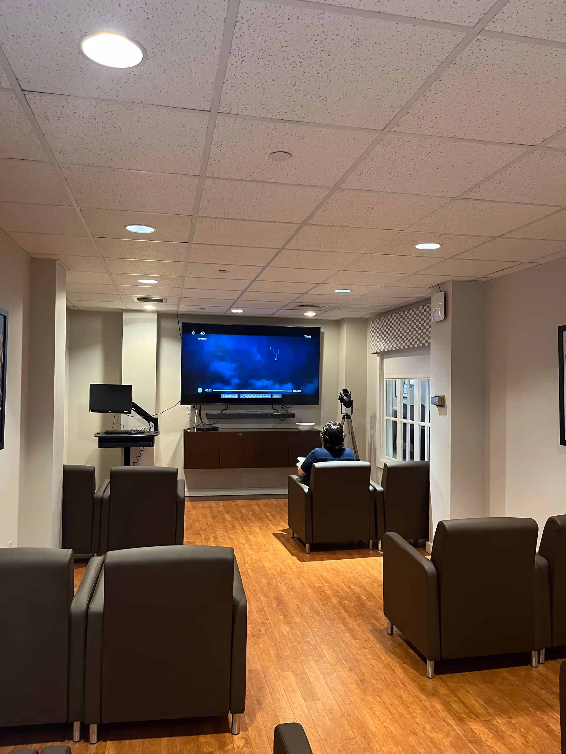 A small lounge area with several brown armchairs arranged facing a large wall-mounted flat screen TV. A person is seated in one of the chairs watching the TV. The room has wood flooring, beige walls, and a drop ceiling with recessed lighting. A window with a patterned valance is visible on the right side.