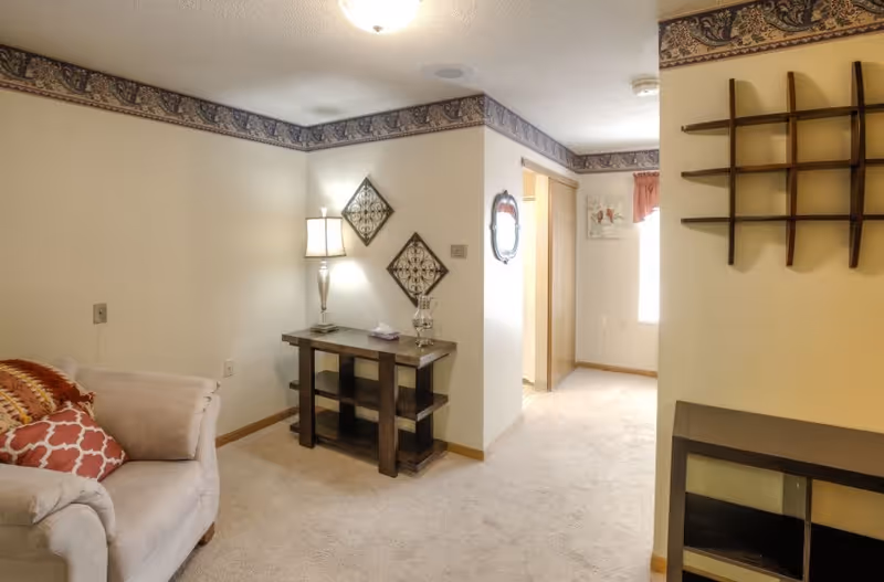 A cozy interior living space featuring a beige armchair with patterned pillows, a wooden side table with a lamp and decorative items, wall-mounted metal art, and a wooden shelving unit. The room has beige carpet, light-colored walls with a decorative border near the ceiling, and a hallway leading to another room with a window and curtains.