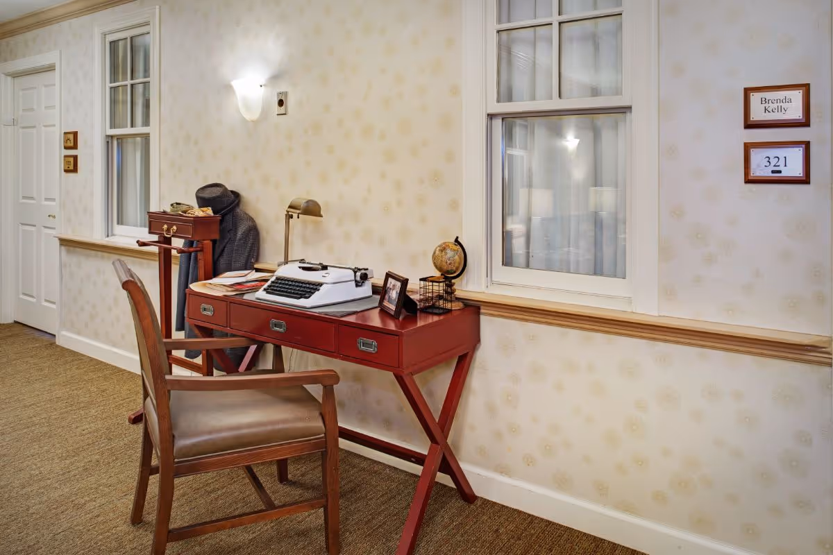 A hallway nook featuring a red writing desk with a typewriter, globe and chair against a wallpapered wall under windows and a room nameplate.