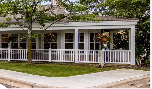 Exterior view of a building with a covered porch featuring white railings and hanging flower baskets, surrounded by green grass and trees.