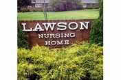 A wooden sign with white letters reading 'LAWSON NURSING HOME' surrounded by green bushes and grass, with a building and windows visible in the background.