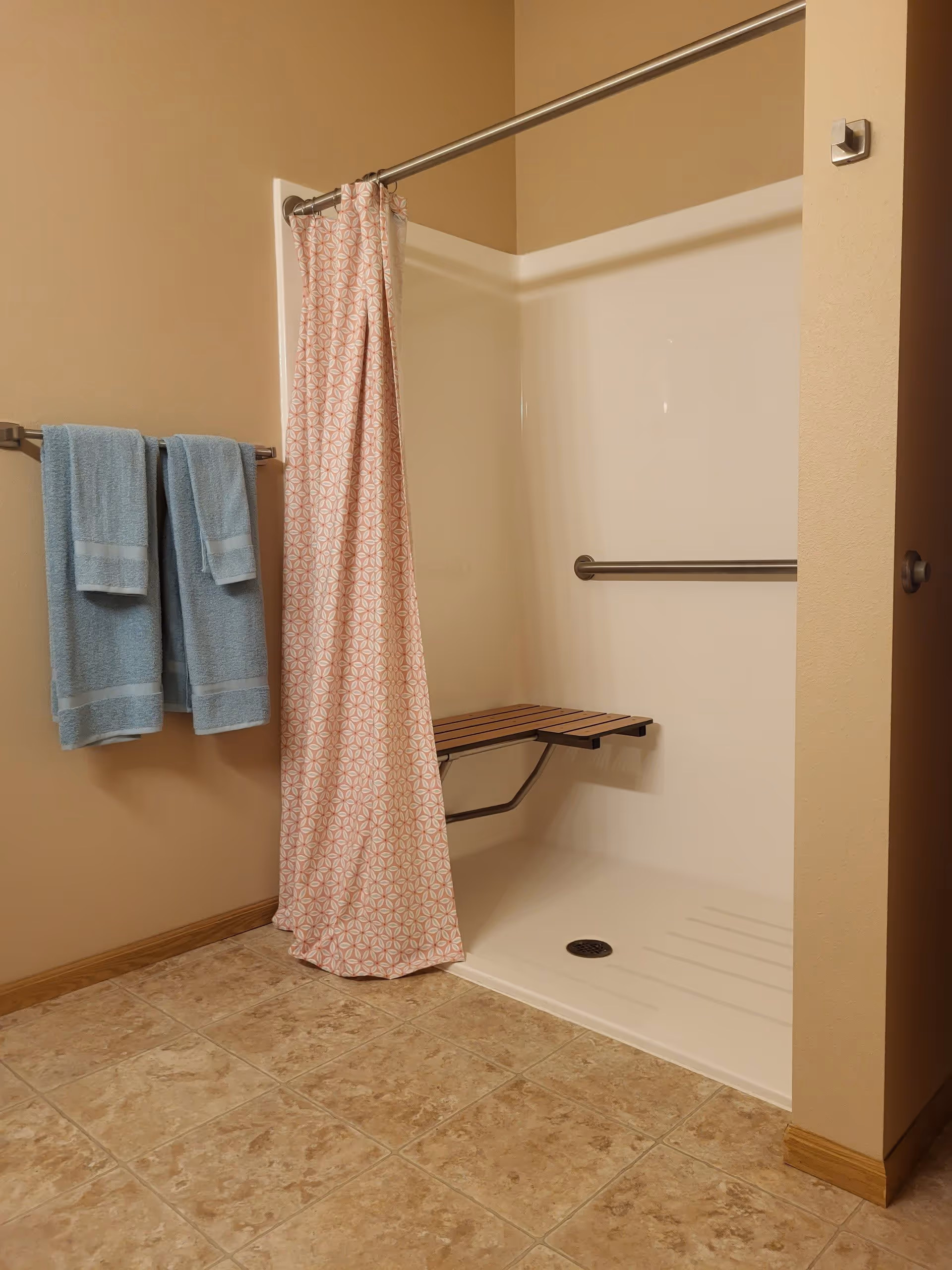 A bathroom shower area with beige walls and tiled floor. The shower has a white base, a metal grab bar, and a foldable wooden shower seat. A pink and white patterned shower curtain hangs from a metal rod. Two blue towels are hanging on a towel rack on the wall next to the shower.