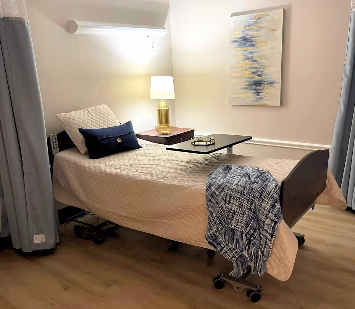 An adjustable hospital bed in a nursing facility room with a bedside table, lamp, overbed tray, decorative pillows, wall art, and a throw blanket.