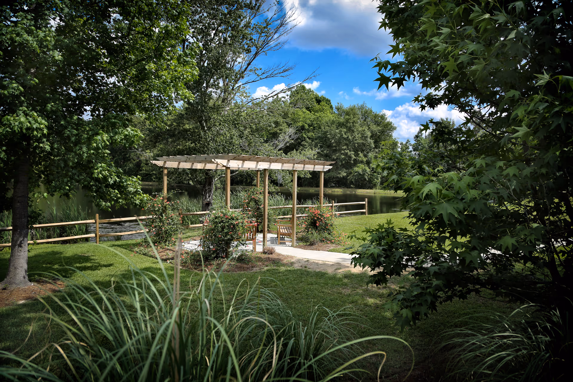 A peaceful outdoor garden area with a wooden pergola surrounded by green trees and bushes. There is a wooden bench under the pergola, a small pathway leading to it, and a wooden fence in the background with a pond and more trees beyond.