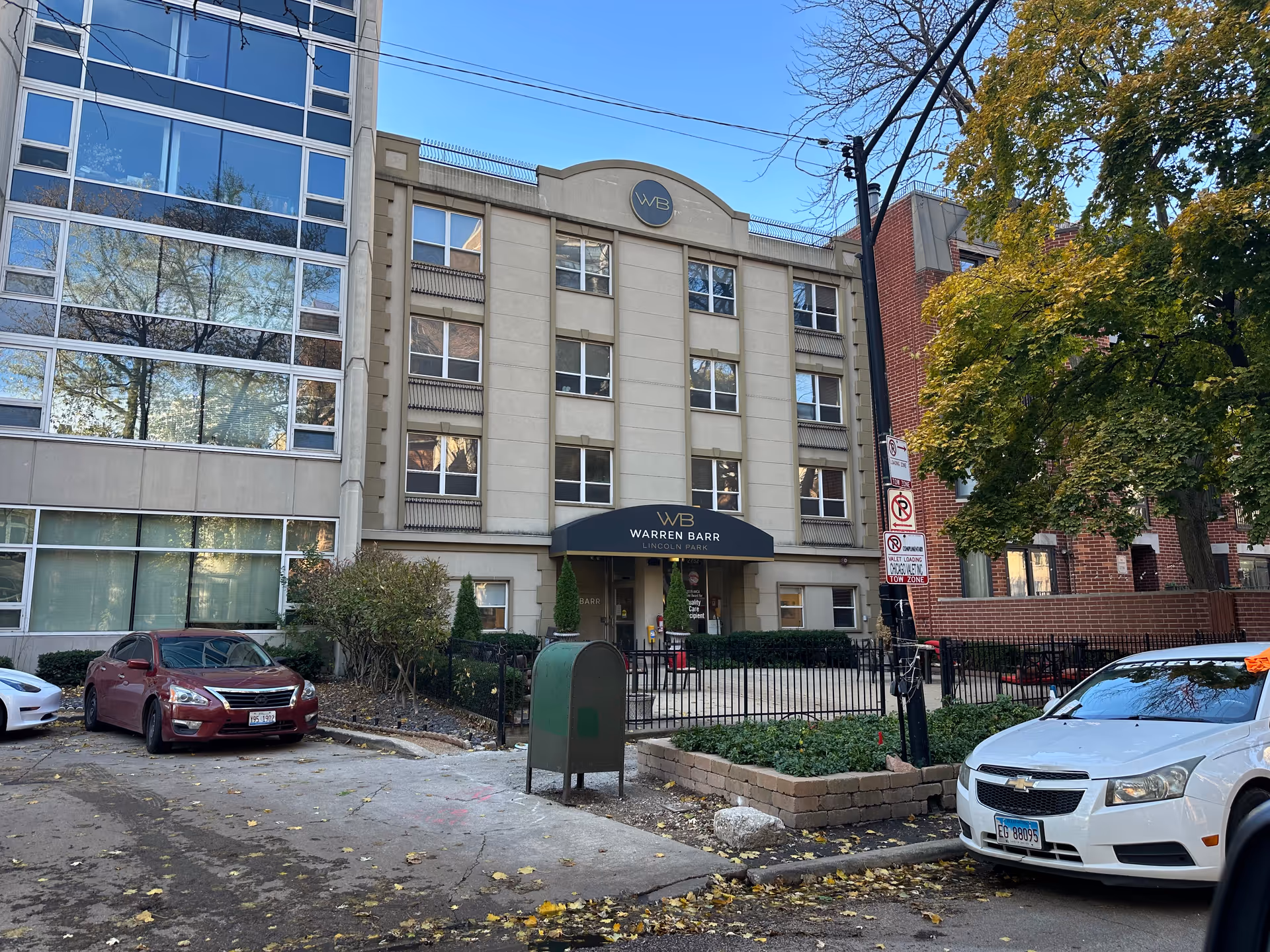 Exterior view of the Warren Barr Lincoln Park senior living facility, showing a multi-story building with a beige facade and a black awning above the entrance displaying the facility name. There are parked cars in front, a green mailbox, and trees with autumn foliage surrounding the area.