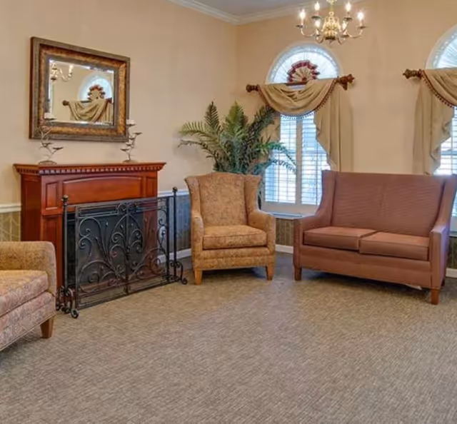 A cozy living room area with beige walls, a carpeted floor, and a decorative fireplace with a black metal screen. There are two upholstered armchairs and a brown loveseat arranged around the fireplace. Two windows with white shutters and beige curtains with valances let in natural light. A chandelier hangs from the ceiling, and a large green plant is placed in the corner.