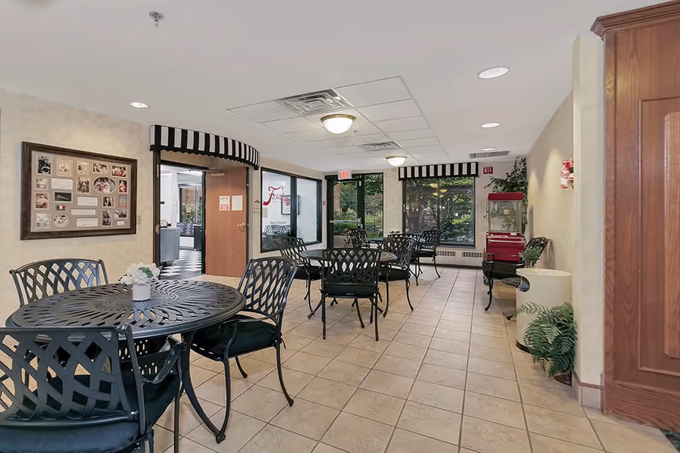 Communal dining area with round metal tables and chairs, tiled floor, and windows overlooking greenery.