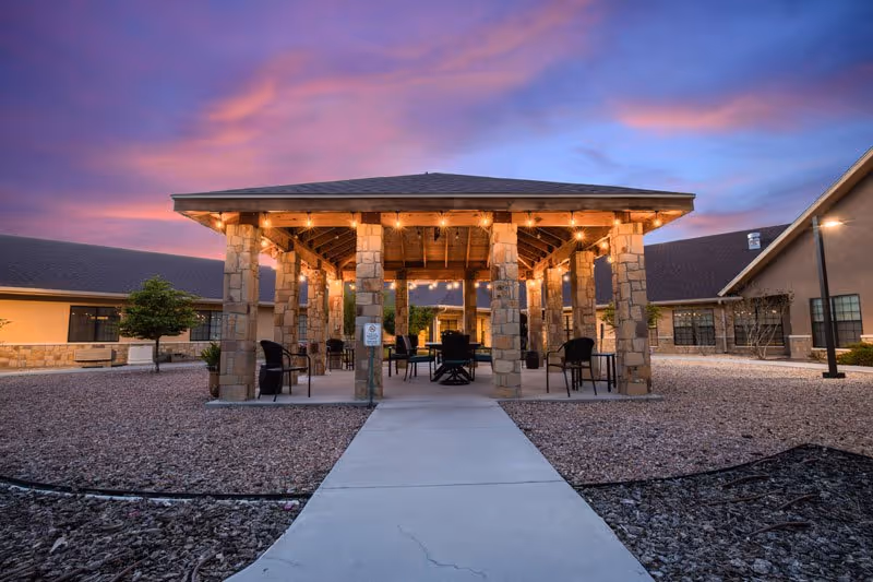 Covered stone pavilion with string lights and seating in a courtyard at sunset.